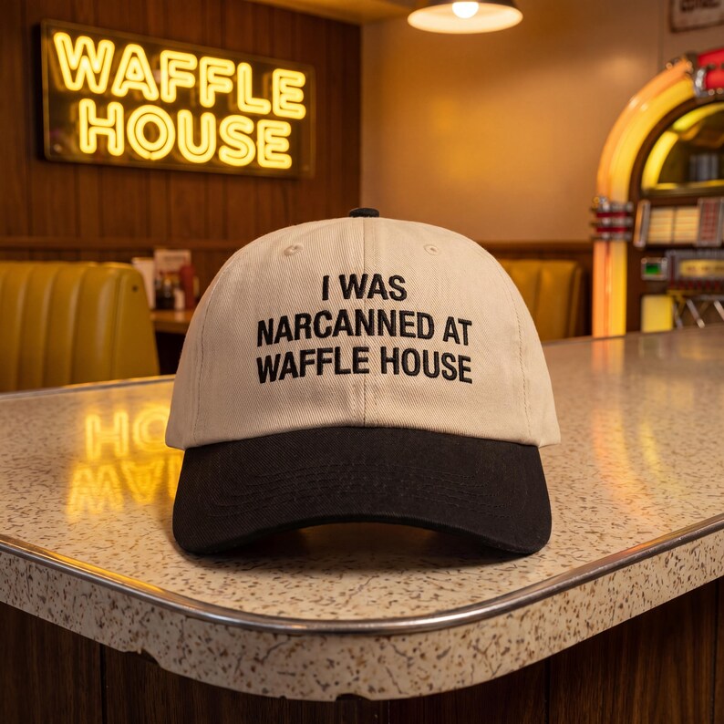 May include: A beige and black baseball cap with the text "I WAS NARCANNED AT WAFFLE HOUSE" in black. The cap is in front of a Waffle House neon sign and a retro diner setting.