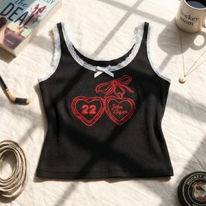 May include: A black ribbed vest top featuring white lace trim along the scoop neckline and a small white bow at the centre. The front displays two red embroidered hearts, one containing the number 22 and the other the text John Logan.