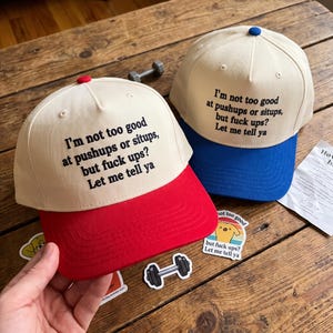 May include: Two baseball caps with off-white crowns and black embroidered text that reads: I'm not too good at pushups or situps, but fuck ups? Let me tell ya. One cap features a red brim and button, while the other has a blue brim and button.