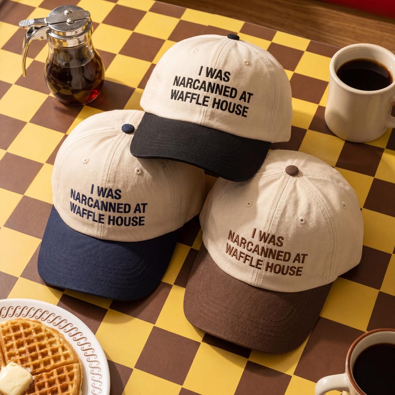 May include: Three beige baseball caps with the text "I WAS NARCANNED AT WAFFLE HOUSE" in black. Each cap has a different coloured brim: black, navy blue, and brown. The caps are arranged on a checkered yellow and brown surface.