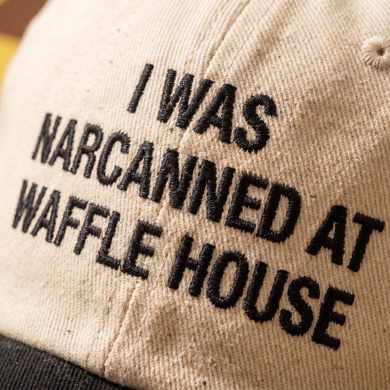 May include: A beige baseball cap with a black peak. The cap has the embroidered words "I WAS NARCANNED AT WAFFLE HOUSE" in black thread. The cap is made of a textured fabric.