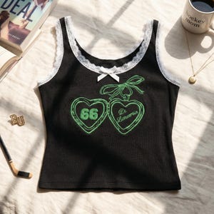May include: A black ribbed tank top featuring white lace trim along the neckline and armholes. A small white bow is centred at the neckline. The front displays two embroidered green hearts with a bow, the number 66, and the text Di. Lanuersin.