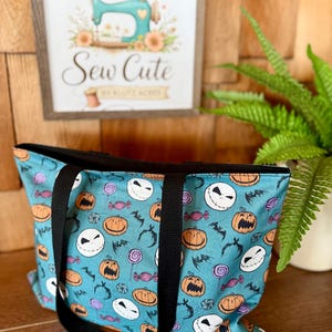 Puede incluir: Un bolso tote turquesa con ribete y asas negras, con un estampado tem&aacute;tico de Halloween de caras de Jack Skellington, calabazas, murci&eacute;lagos y caramelos. La bolsa se muestra frente a un fondo de madera y un letrero enmarcado que dice "Sew Cute".