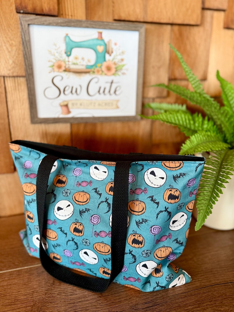 Puede incluir: Un bolso tote turquesa con ribete y asas negras, con un estampado tem&aacute;tico de Halloween de caras de Jack Skellington, calabazas, murci&eacute;lagos y caramelos. La bolsa se muestra frente a un fondo de madera y un letrero enmarcado que dice "Sew Cute".