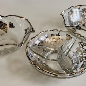May include: Three clear glass serving bowls with silver rims and floral designs. The bowls have a scalloped edge. One bowl is divided into three sections. The floral design includes silver and gold accents.