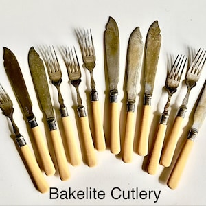 May include: A collection of vintage Bakelite cutlery, including forks and knives. The knives have a curved blade design. The handles are a light cream color, and the metal is tarnished. The text "Bakelite Cutlery" is at the bottom.