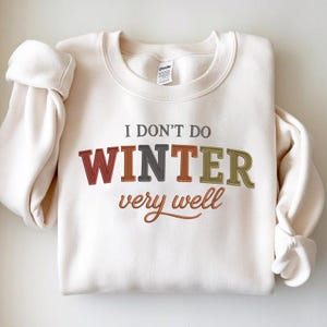 May include: Cream-colored sweatshirt with the text "I DON'T DO WINTER very well" in a mix of colors. The word "WINTER" is in a bold, multi-colored font, with the sleeves folded.