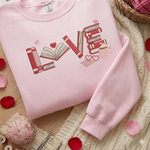 May include: A light pink sweatshirt with the word "LOVE" embroidered in red. The "L" is a stack of books, the "O" is an open book with a heart, the "V" is a book, and the "E" is a stack of books with a mug and hearts.