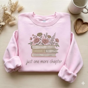 May include: A light pink sweatshirt with a floral and book design. The graphic features a stack of books topped with flowers and the phrase "just one more chapter" in a script font. The sweatshirt has a crew neck and long sleeves.