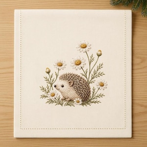 May include: A square off-white fabric napkin featuring an embroidered design of a small brown hedgehog surrounded by white daisies with yellow centers and green stems. The napkin has a decorative stitched border along its four edges.