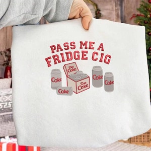 May include: White sweatshirt with a red graphic that says "PASS ME A FRIDGE CIG". The graphic includes illustrations of Coke and Diet Coke cans and a six-pack of Diet Coke.