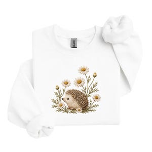 May include: A white long-sleeve sweatshirt featuring a central embroidered graphic of a small brown hedgehog surrounded by several white and yellow daisy flowers with green stems and leaves. The garment has a crew neckline and ribbed cuffs.