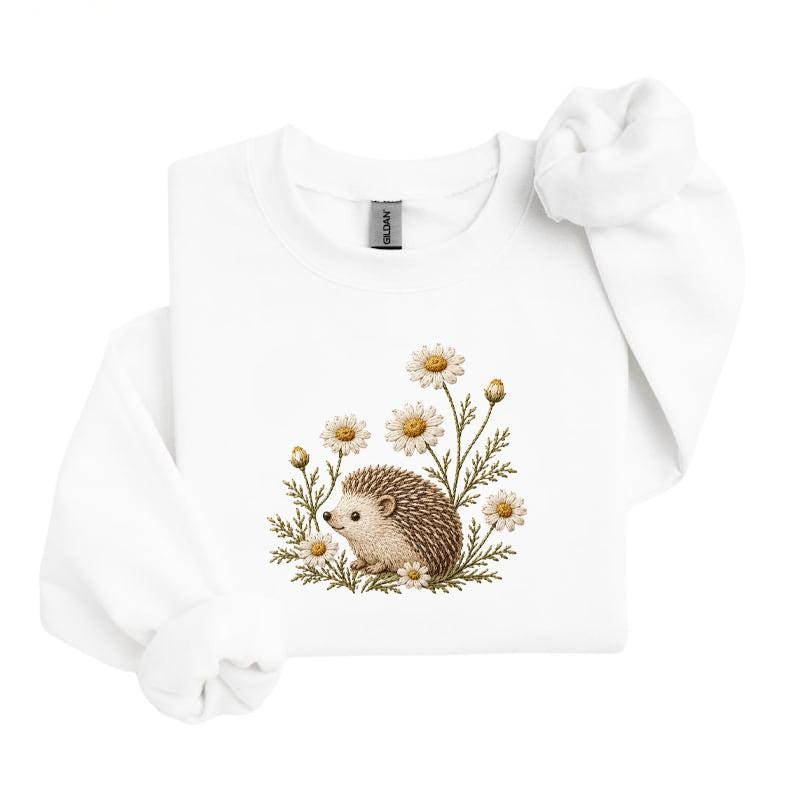 May include: A white long-sleeve sweatshirt featuring a central embroidered graphic of a small brown hedgehog surrounded by several white and yellow daisy flowers with green stems and leaves. The garment has a crew neckline and ribbed cuffs.