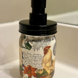 May include: A glass soap dispenser featuring a black plastic pump and lid. The jar is decorated with a Farmers Market graphic, a rooster, a watering can, orange flowers, and cursive script text. The design has a cream-colored background.