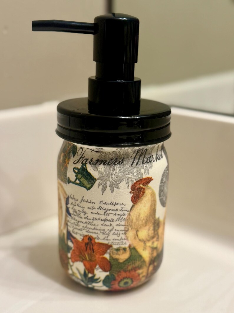 May include: A glass soap dispenser featuring a black plastic pump and lid. The jar is decorated with a Farmers Market graphic, a rooster, a watering can, orange flowers, and cursive script text. The design has a cream-colored background.