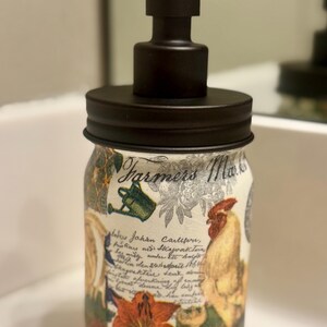 May include: A glass soap dispenser featuring a matte black pump and lid. The jar is decorated with a collage of images including a rooster, a watering can, flowers, and cursive script text that reads Farmers Market and Johan Carlsen.
