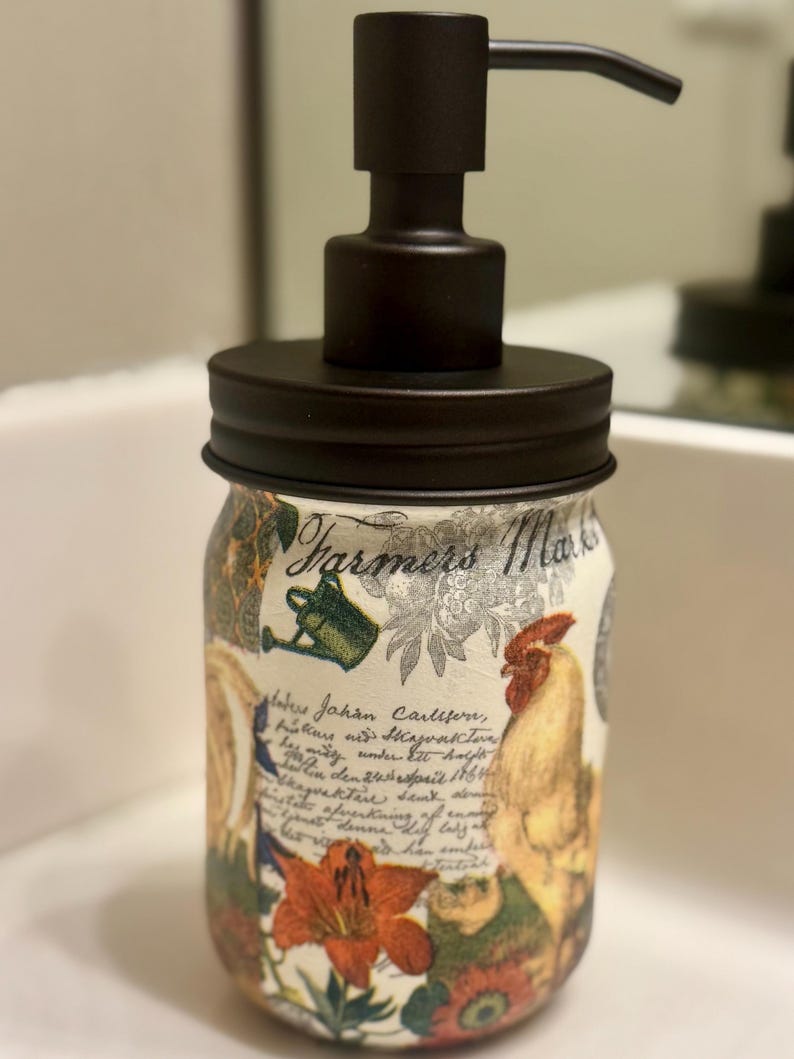 May include: A glass soap dispenser featuring a matte black pump and lid. The jar is decorated with a collage of images including a rooster, a watering can, flowers, and cursive script text that reads Farmers Market and Johan Carlsen.