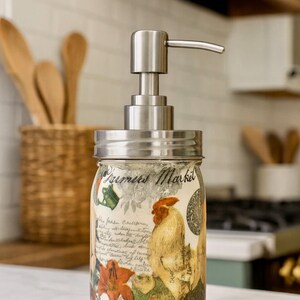 May include: A glass soap dispenser featuring a brushed metal pump and a screw-top lid. The jar is decorated with a colorful rooster illustration, orange flowers, and the text Farmers Market written in a cursive script over handwritten style notes.