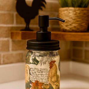 May include: A glass soap dispenser with a matte black pump and lid. The jar features a Farmers Market label, a rooster illustration, floral motifs, and cursive script text. The design includes warm tones of orange, yellow, and green on a light background.