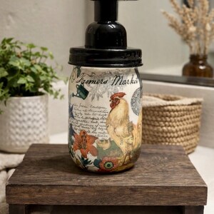 May include: A glass soap dispenser with a black pump lid featuring a decorative wrap with the text Farmers Market, a rooster illustration, floral patterns, and script writing. The jar has a rounded shape and a cream-colored background.