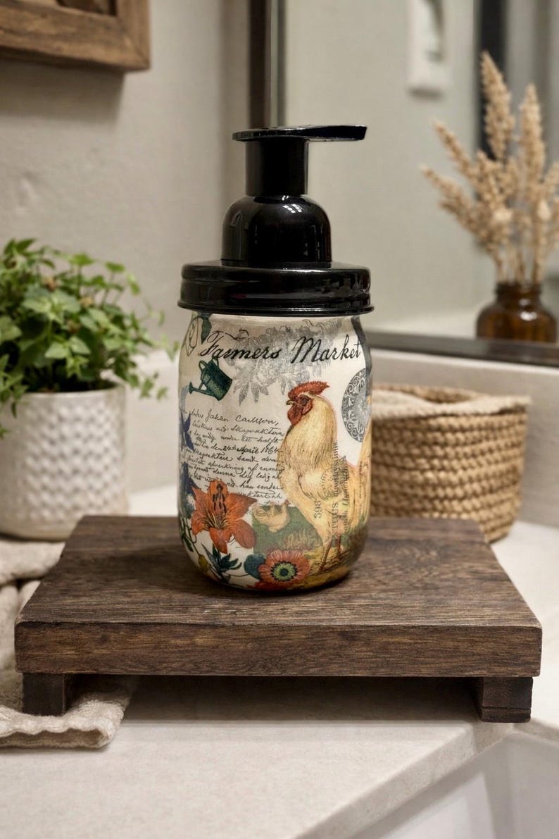 May include: A glass soap dispenser with a black pump lid featuring a decorative wrap with the text Farmers Market, a rooster illustration, floral patterns, and script writing. The jar has a rounded shape and a cream-colored background.
