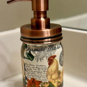 May include: A soap dispenser featuring a copper-colored pump and lid atop a glass jar. The jar is decorated with a Farmers Market label, a rooster illustration, floral motifs, and cursive script text in black ink over a cream-colored background.