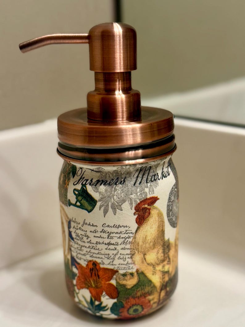 May include: A soap dispenser featuring a copper-colored pump and lid atop a glass jar. The jar is decorated with a Farmers Market label, a rooster illustration, floral motifs, and cursive script text in black ink over a cream-colored background.