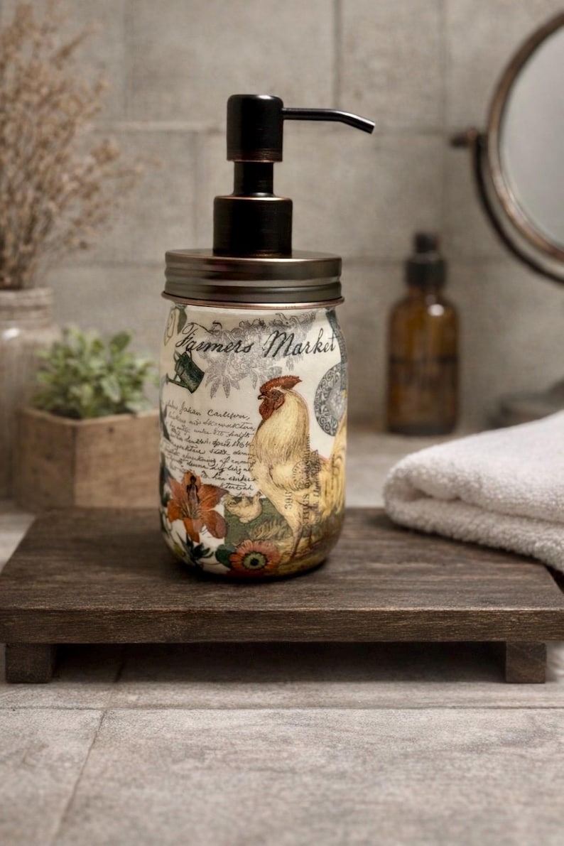 May include: A glass soap dispenser featuring a dark bronze pump and lid. The jar is decorated with a vintage-style graphic print including a rooster, floral elements, and the text Farmers Market in a cursive script, along with various botanical illustrations.