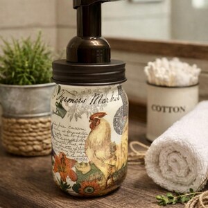 May include: A glass soap dispenser with a dark brown pump and lid. The jar features a decorative print with the text Farmers Market, a rooster illustration, floral motifs, and cursive script text in shades of orange, yellow, and green.