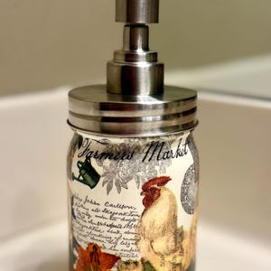 May include: A soap dispenser featuring a glass jar base with a brushed metal pump lid. The jar is decorated with a Farmers Market print, a rooster illustration, floral motifs, and cursive script text. The design includes green, red, and orange colors.
