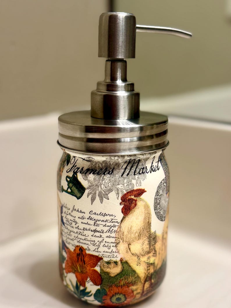May include: A soap dispenser featuring a glass jar base with a brushed metal pump lid. The jar is decorated with a Farmers Market print, a rooster illustration, floral motifs, and cursive script text. The design includes green, red, and orange colors.