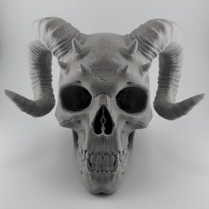 May include: A gray skull sculpture with large curved horns and smaller horns above the eye sockets. The skull has detailed features, including eye sockets, a nasal cavity, and teeth. The sculpture is set against a plain white background.