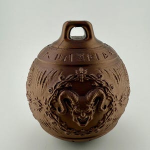 Puede incluir: Una campana esférica de color bronce con un lazo en la parte superior. La campana presenta intrincados diseños en relieve, incluida una cabeza de carnero dentro de una corona floral. La superficie está texturizada con líneas verticales y caracteres asiáticos.