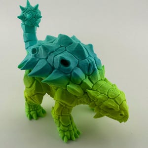 May include: A 3D-printed model of an Ankylosaurus dinosaur. The model features a gradient of colors, transitioning from green to blue. The dinosaur has a spiky tail club and armored plates.