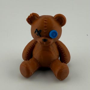 May include: A brown teddy bear figurine with a blue eye and an X over the other eye. The bear has stitched details and a small tear on one ear. The bear is sitting with its legs extended.