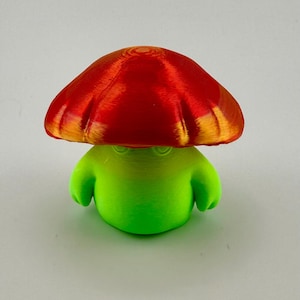 May include: A 3D-printed mushroom figurine with a bright red cap and a green body. The cap has a textured surface with orange highlights. The figurine has a whimsical, cartoonish design.