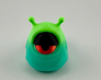 May include: A 3D-printed, cartoonish creature with a large, single eye. The body transitions from green to teal, with a black eye and a red iris. Two small antennae extend from the top of the head. The design is smooth and rounded.