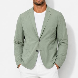 Puede incluir: Un blazer de color verde salvia con solapa de muesca, dos botones oscuros para el cierre y dos bolsillos delanteros de parche. El blazer se lleva sobre una camiseta blanca lisa, mostrando un acabado de tela texturizada con mangas largas.