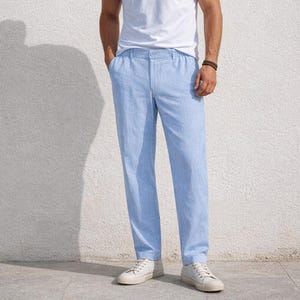 May include: Light blue linen pants with a relaxed fit. The pants have a button and zipper closure, and pockets. The model is wearing white sneakers and a white t-shirt. The pants are made of a lightweight fabric.