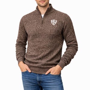 Könnte beinhalten: Brauner Strickpullover mit einem Kragen mit Viertelreißverschluss und einem weißen Emblem auf der Brust. Der Pullover hat ein strukturiertes Muster und lange Ärmel. Die Person trägt eine blaue Jeans.