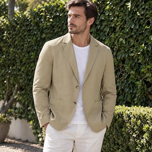 Puede incluir: Una americana de color beige con solapa de muesca y dos botones frontales, combinada con una camiseta blanca lisa y pantalones claros. La prenda cuenta con mangas largas y dos bolsillos de parche inferiores, ofreciendo un diseño sencillo.