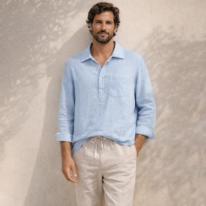 Puede incluir: Una camisa de lino azul claro con mangas largas y cuello abotonado. La camisa tiene un bolsillo en el pecho y se combina con pantalones de lino beige con cordón. El modelo está de pie frente a una pared de tonos neutros.