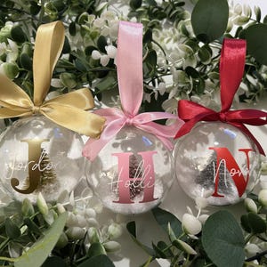 May include: Three clear baubles with personalised names and initial letters. Each bauble has a different coloured ribbon: gold, pink, and red. The baubles are filled with faux snow and a small Christmas tree.