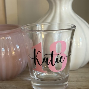 May include: A clear shot glass featuring the name "Katie" in black script and the number "18" in pink. The glass is set on a wooden surface, with a pink candle holder and a white vase behind it.