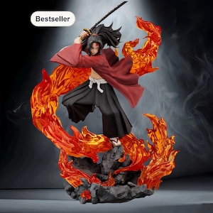Op de afbeelding: Gedetailleerd beeldje van een personage in een dynamische pose, met een katana. Het figuur draagt een rode mantel en een zwarte broek, met lang zwart haar. De basis heeft vurige oranje en rode vlammen. Het woord "Bestseller" staat in een witte ovaal.