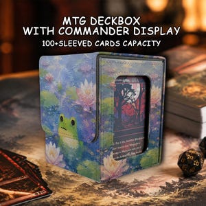 Puede incluir: Una caja para barajas MTG rectangular con ventana para comandante, con un estampado de una rana verde entre nenúfares rosas. El texto dice MTG DECKBOX WITH COMMANDER DISPLAY y 100+ SLEEVED CARDS CAPACITY. La caja tiene un acabado texturizado.