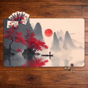 Könnte beinhalten: Gaming-Matte mit einer asiatisch inspirierten Landschaft mit roter Sonne, Bergen und einem Baum mit roten Blättern. Ein kleines Boot mit einer Person befindet sich auf dem Wasser. Inklusive passender Kartenhüllen und Würfel.