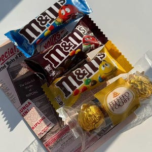 Könnte beinhalten: Eine Auswahl an M&M's Schokoriegeln in blauen, braunen und gelben Verpackungen, zusammen mit einer goldverpackten Ferrero Rocher Praline. Ein Vintage-Zeitungsartikel mit dem Text "KIPLING" ist ebenfalls zu sehen.