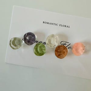 Könnte beinhalten: Sechs durchscheinende, runde Perlen in verschiedenen Farben: Grün, Lila, Klar, Braun und Rosa. Die Perlen sind auf einer weißen Karte mit dem Text "ROMANTIC FLORAL" angeordnet.