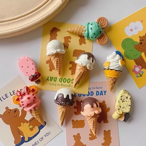May include: A collection of miniature ice cream cone and popsicle ornaments featuring various flavors, colors, and toppings. The set includes detailed waffle cones, chocolate drizzles, cookie ears, and fruit accents, arranged on decorative cards.
